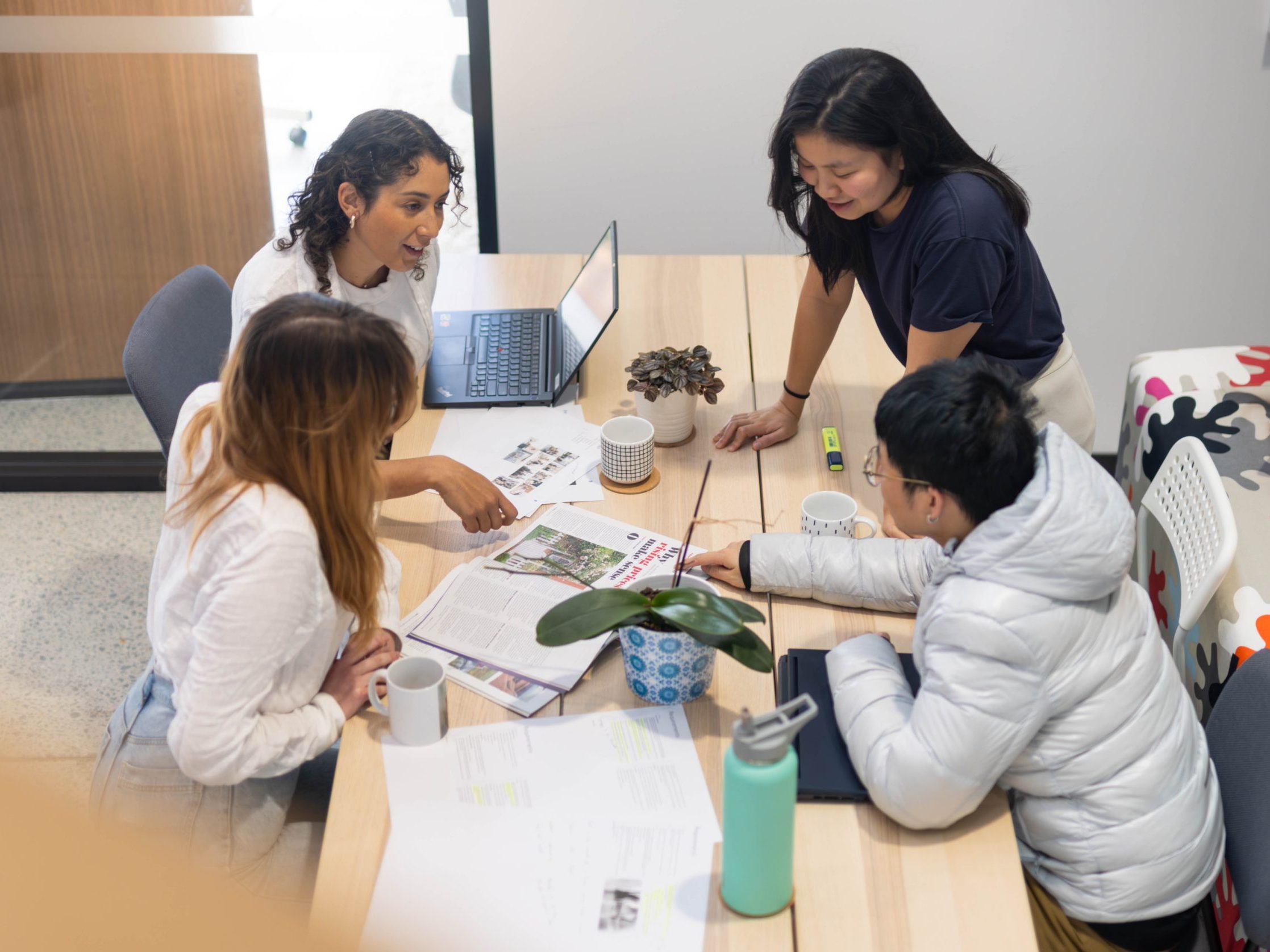 Four people engage in a meeting around a table, surrounded by documents, a laptop, coffee cups, and a plant, as they discuss key elements of copywriting in an office setting.