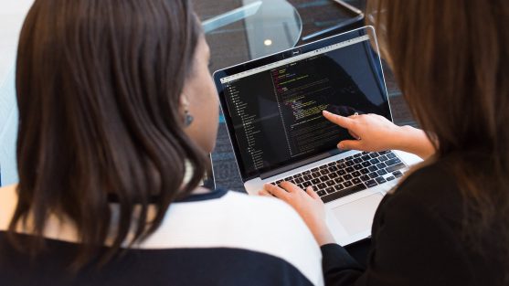 Two people are examining code on a laptop screen, with one pointing at the display.