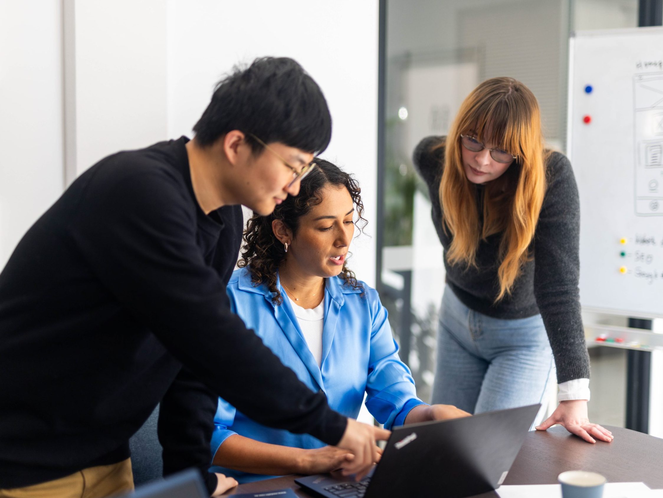 Three people collaborate around a laptop in an office setting, discussing WordPress maintenance strategies, with a whiteboard filled with ideas in the background.