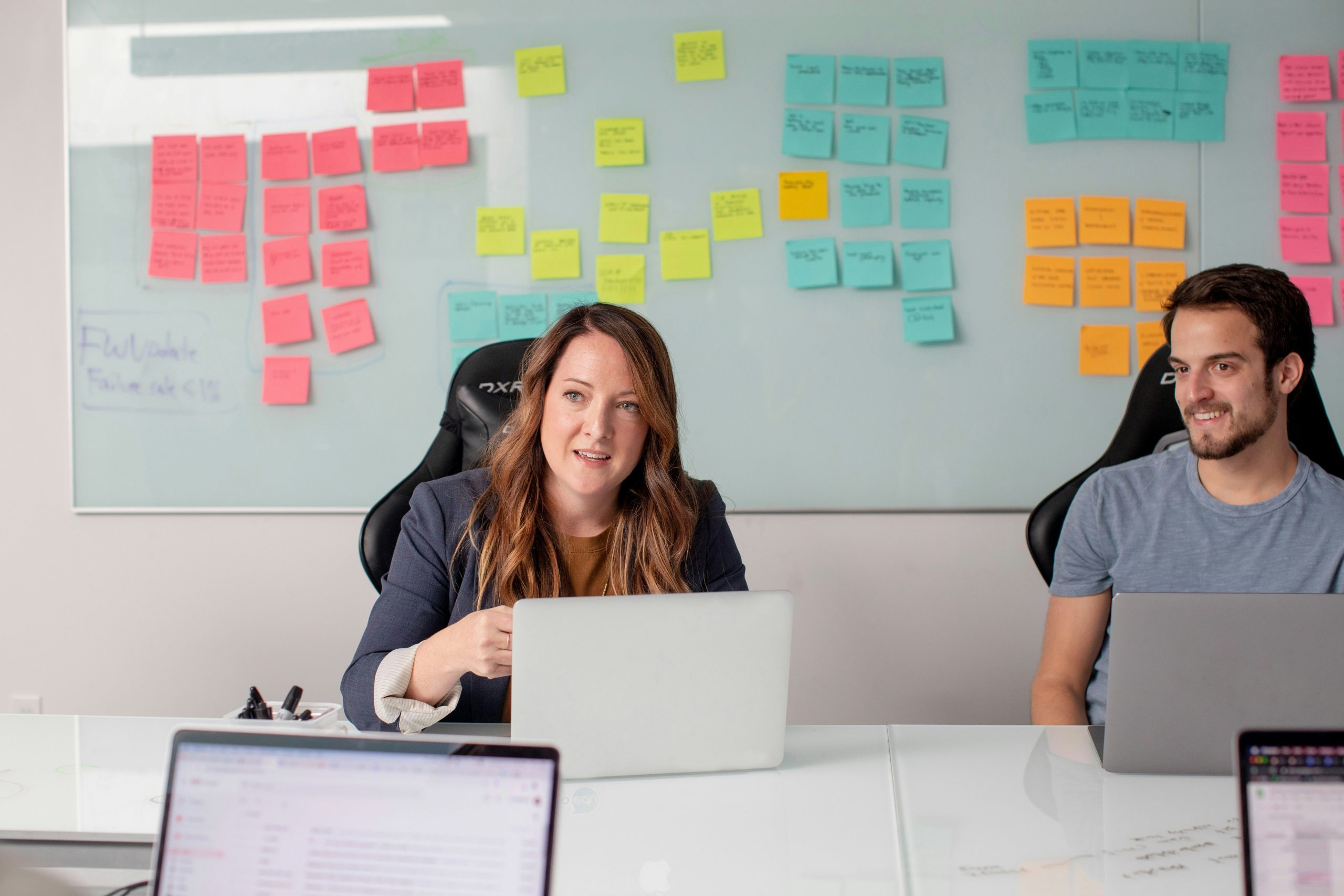 A man and woman in a meeting discussing plans to launch a website. In the background, there is a whiteboard with different coloured post it notes.