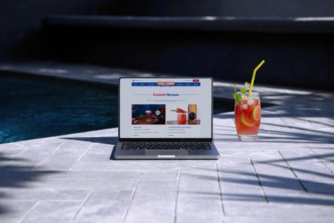 A poolside laptop beside a cocktail. The laptop shows the Local Liquor website.