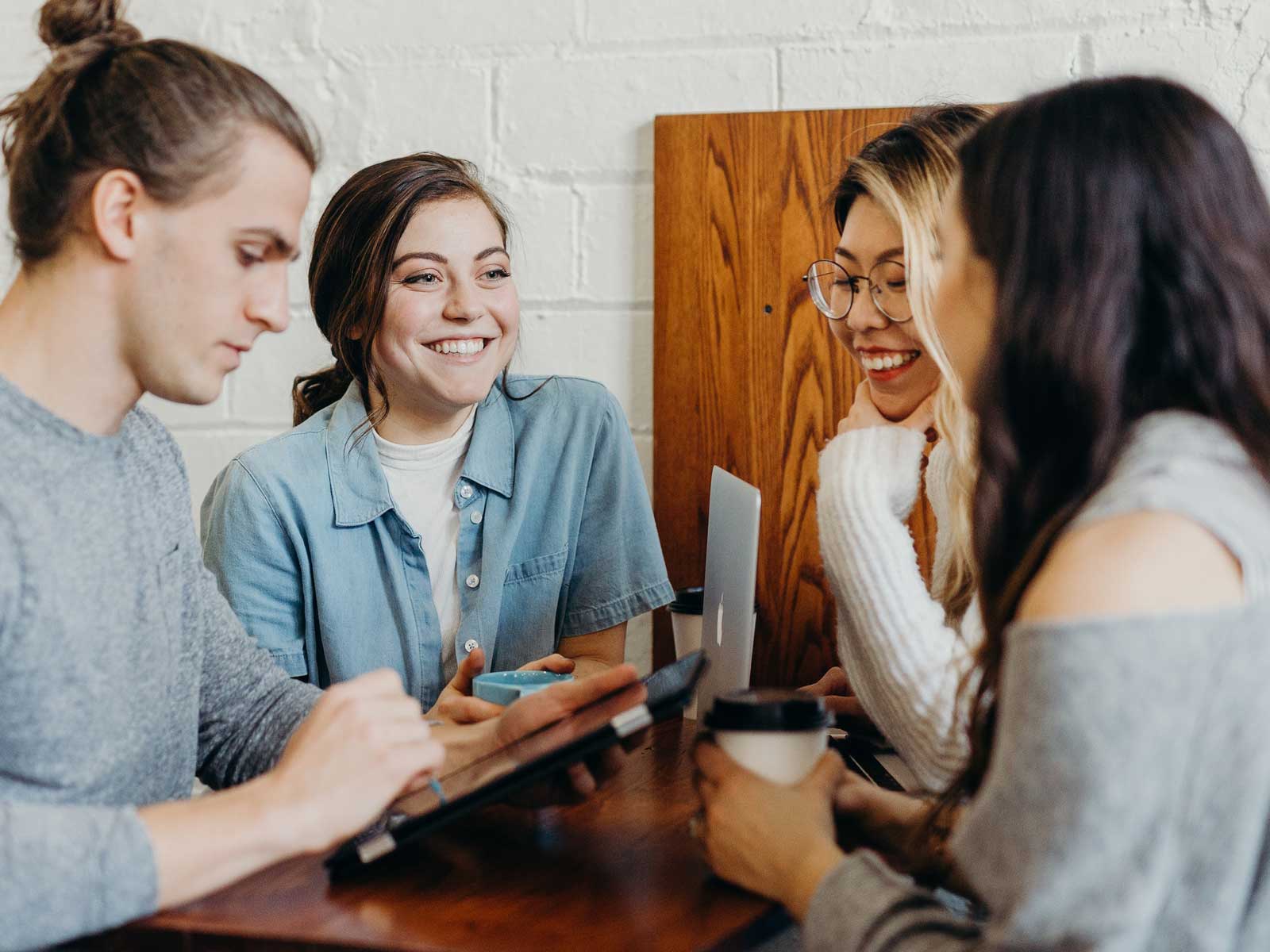Four people sitting at a table in a casual setting, engaged in conversation about the latest trends in web design. Two have open laptops displaying colorful interfaces, and one is holding a coffee cup.