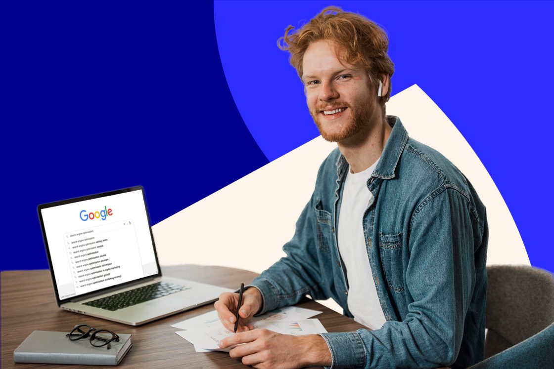 A man with red hair and a beard, wearing a denim jacket over a white shirt, sits at a desk in Canberra with a laptop showing an SEO-related Google search and papers. He is smiling and holding a pen.